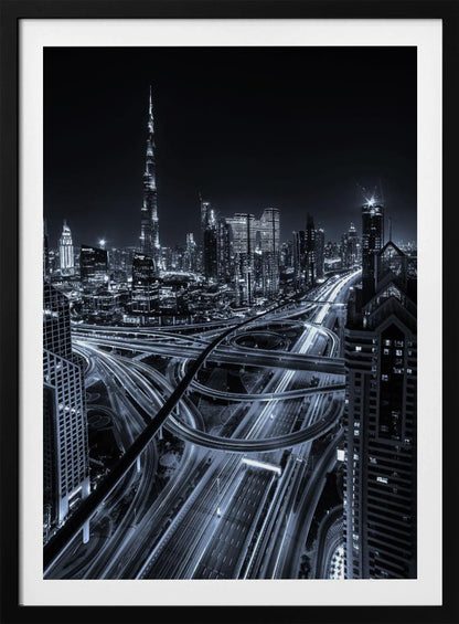 A cool-toned black and white photograph of the Dubai skyline at night. The iconic Burj Khalifa stands tall in the background, while the foreground is dominated by a massive, complex highway interchange with light trails from moving cars, creating a sense of motion and modernity. Poster