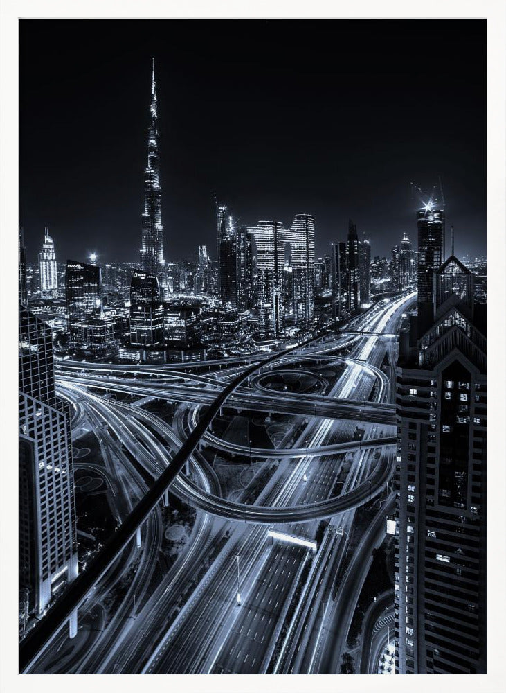 A cool-toned black and white photograph of the Dubai skyline at night. The iconic Burj Khalifa stands tall in the background, while the foreground is dominated by a massive, complex highway interchange with light trails from moving cars, creating a sense of motion and modernity. Poster