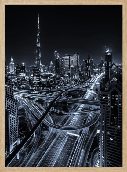 A cool-toned black and white photograph of the Dubai skyline at night. The iconic Burj Khalifa stands tall in the background, while the foreground is dominated by a massive, complex highway interchange with light trails from moving cars, creating a sense of motion and modernity. Poster