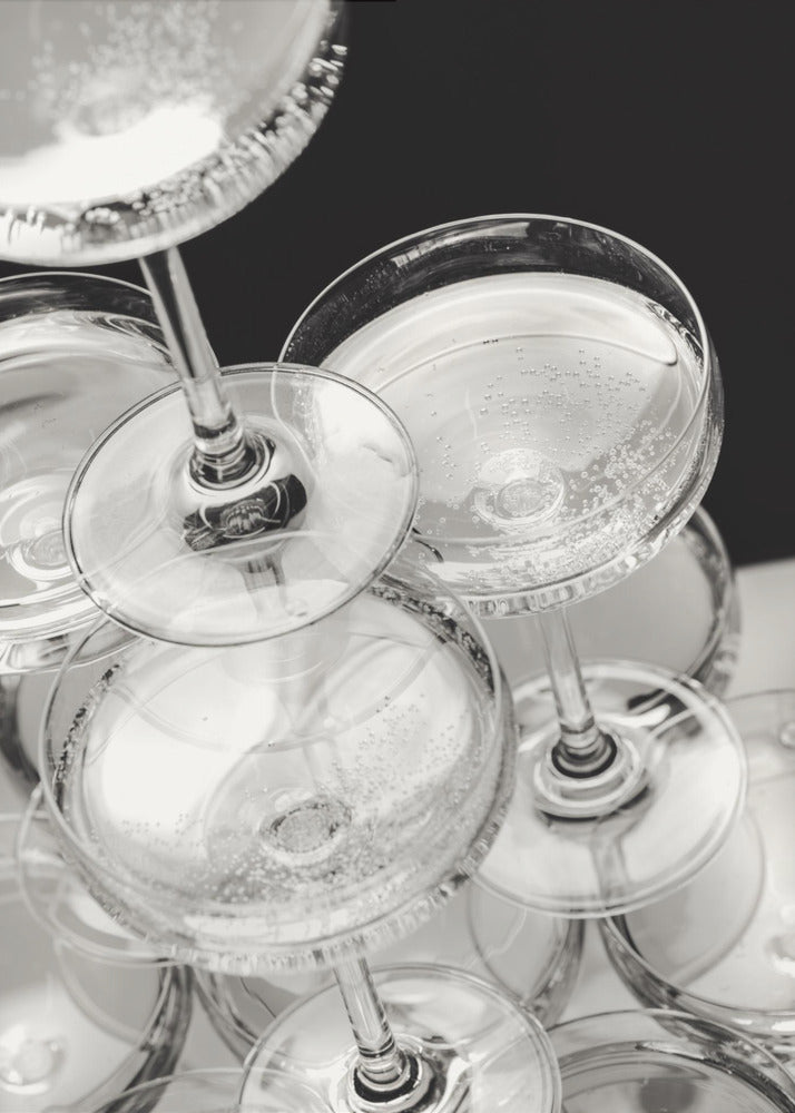 A close-up, high-angle photograph in black and white of a champagne tower. Several coupe-style glasses are stacked, filled with bubbly champagne that catches the light. The background is dark, creating a strong contrast and highlighting the elegant glassware. Decor