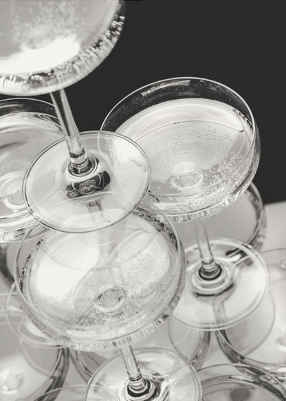 A close-up, high-angle photograph in black and white of a champagne tower. Several coupe-style glasses are stacked, filled with bubbly champagne that catches the light. The background is dark, creating a strong contrast and highlighting the elegant glassware. Decor