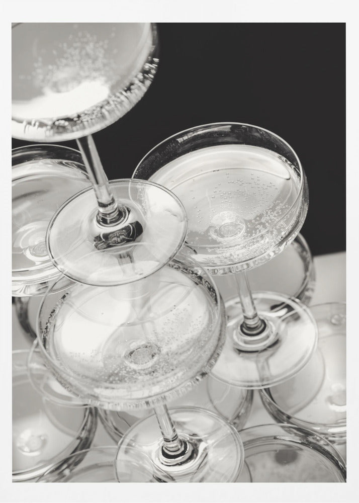 A close-up, high-angle photograph in black and white of a champagne tower. Several coupe-style glasses are stacked, filled with bubbly champagne that catches the light. The background is dark, creating a strong contrast and highlighting the elegant glassware. Decor