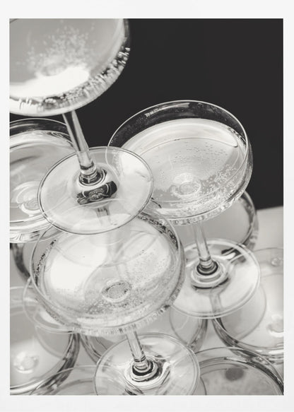A close-up, high-angle photograph in black and white of a champagne tower. Several coupe-style glasses are stacked, filled with bubbly champagne that catches the light. The background is dark, creating a strong contrast and highlighting the elegant glassware. Decor