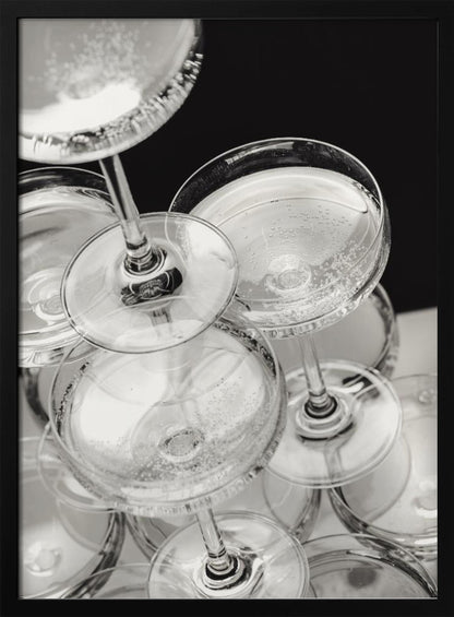 A close-up, high-angle photograph in black and white of a champagne tower. Several coupe-style glasses are stacked, filled with bubbly champagne that catches the light. The background is dark, creating a strong contrast and highlighting the elegant glassware. Decor