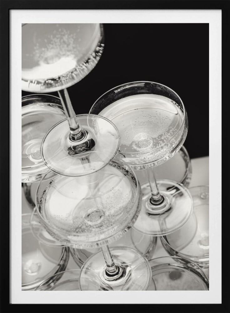 A close-up, high-angle photograph in black and white of a champagne tower. Several coupe-style glasses are stacked, filled with bubbly champagne that catches the light. The background is dark, creating a strong contrast and highlighting the elegant glassware. Decor
