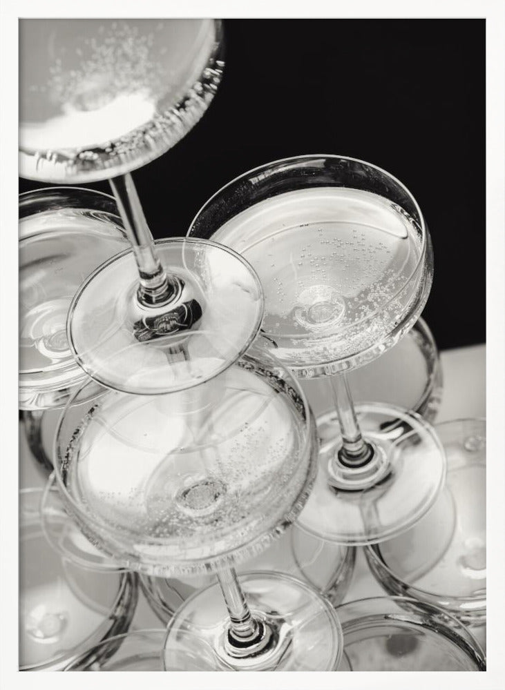 A close-up, high-angle photograph in black and white of a champagne tower. Several coupe-style glasses are stacked, filled with bubbly champagne that catches the light. The background is dark, creating a strong contrast and highlighting the elegant glassware. Decor