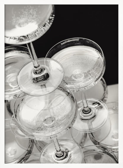A close-up, high-angle photograph in black and white of a champagne tower. Several coupe-style glasses are stacked, filled with bubbly champagne that catches the light. The background is dark, creating a strong contrast and highlighting the elegant glassware. Decor