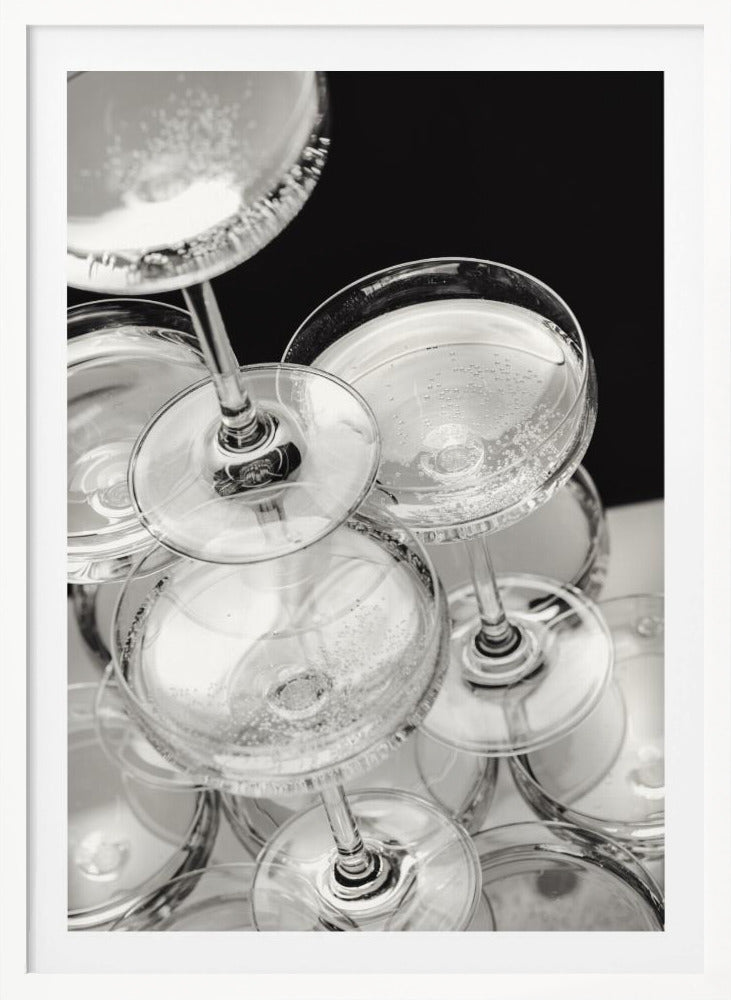 A close-up, high-angle photograph in black and white of a champagne tower. Several coupe-style glasses are stacked, filled with bubbly champagne that catches the light. The background is dark, creating a strong contrast and highlighting the elegant glassware. Decor