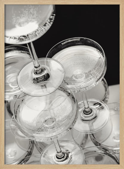 A close-up, high-angle photograph in black and white of a champagne tower. Several coupe-style glasses are stacked, filled with bubbly champagne that catches the light. The background is dark, creating a strong contrast and highlighting the elegant glassware. Decor