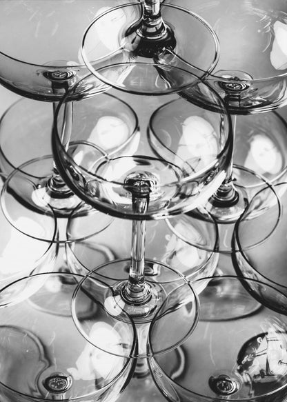A black and white, overhead photograph of a tightly packed stack of empty champagne coupe glasses. The overlapping rims, stems, and bowls create a complex abstract pattern of circles and reflections, all enclosed within a simple black frame. Poster