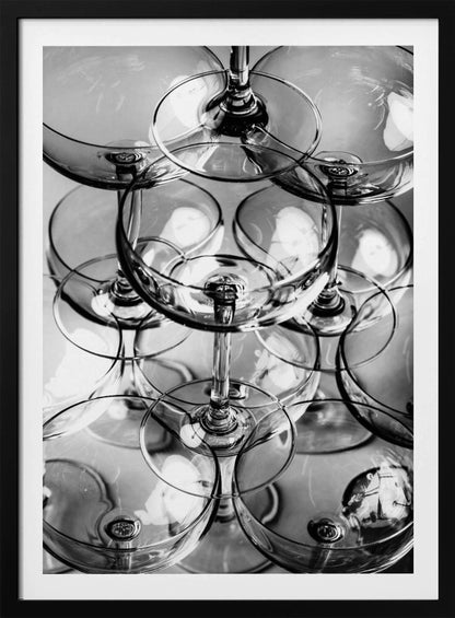 A black and white, overhead photograph of a tightly packed stack of empty champagne coupe glasses. The overlapping rims, stems, and bowls create a complex abstract pattern of circles and reflections, all enclosed within a simple black frame. Poster