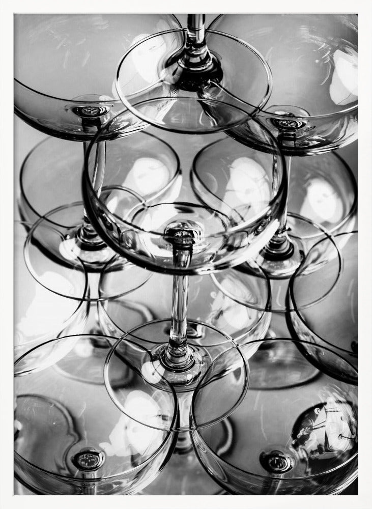 A black and white, overhead photograph of a tightly packed stack of empty champagne coupe glasses. The overlapping rims, stems, and bowls create a complex abstract pattern of circles and reflections, all enclosed within a simple black frame. Poster