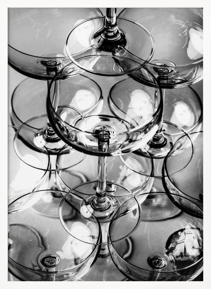 A black and white, overhead photograph of a tightly packed stack of empty champagne coupe glasses. The overlapping rims, stems, and bowls create a complex abstract pattern of circles and reflections, all enclosed within a simple black frame. Poster