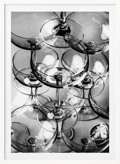 A black and white, overhead photograph of a tightly packed stack of empty champagne coupe glasses. The overlapping rims, stems, and bowls create a complex abstract pattern of circles and reflections, all enclosed within a simple black frame. Poster