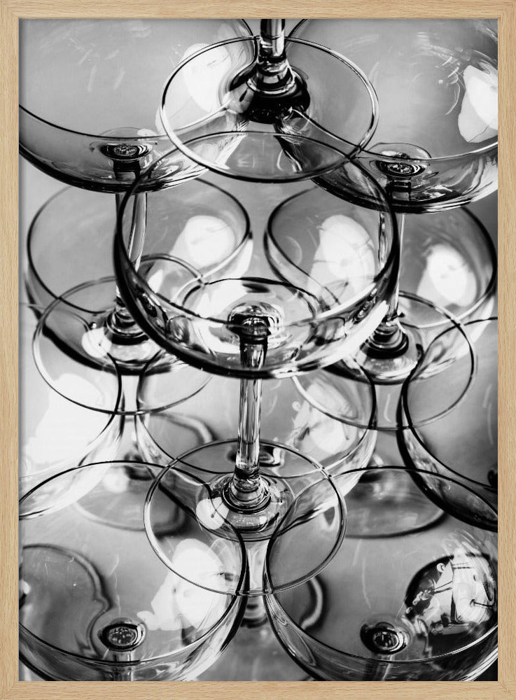 A black and white, overhead photograph of a tightly packed stack of empty champagne coupe glasses. The overlapping rims, stems, and bowls create a complex abstract pattern of circles and reflections, all enclosed within a simple black frame. Poster