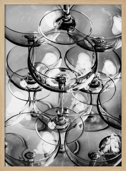 A black and white, overhead photograph of a tightly packed stack of empty champagne coupe glasses. The overlapping rims, stems, and bowls create a complex abstract pattern of circles and reflections, all enclosed within a simple black frame. Poster