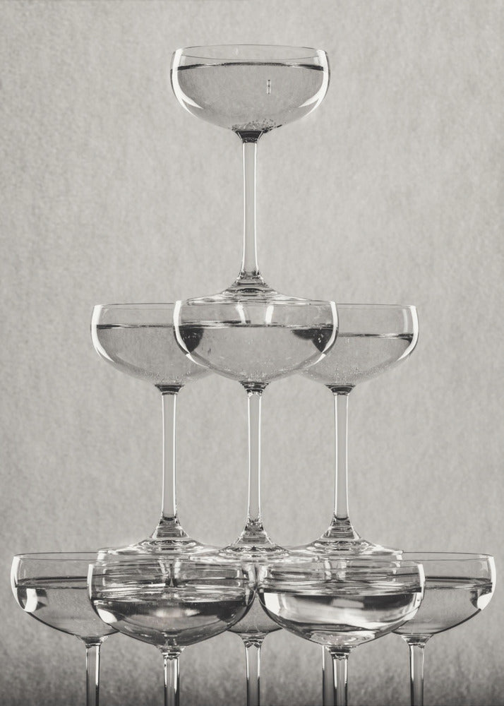 A classic black and white photograph of a champagne tower, with coupe glasses stacked in a pyramid. The glasses are filled with bubbly liquid, and the composition is set against a textured light gray background and enclosed in a black frame. Poster