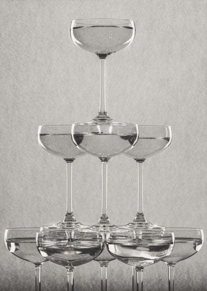 A classic black and white photograph of a champagne tower, with coupe glasses stacked in a pyramid. The glasses are filled with bubbly liquid, and the composition is set against a textured light gray background and enclosed in a black frame. Poster