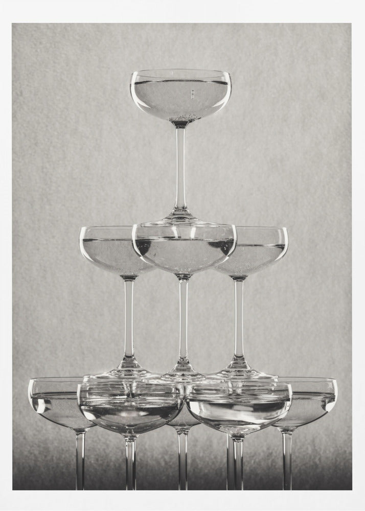 A classic black and white photograph of a champagne tower, with coupe glasses stacked in a pyramid. The glasses are filled with bubbly liquid, and the composition is set against a textured light gray background and enclosed in a black frame. Poster