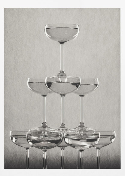 A classic black and white photograph of a champagne tower, with coupe glasses stacked in a pyramid. The glasses are filled with bubbly liquid, and the composition is set against a textured light gray background and enclosed in a black frame. Poster