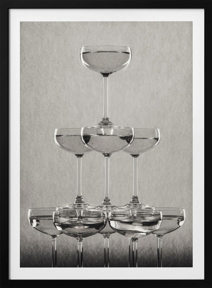 A classic black and white photograph of a champagne tower, with coupe glasses stacked in a pyramid. The glasses are filled with bubbly liquid, and the composition is set against a textured light gray background and enclosed in a black frame. Poster