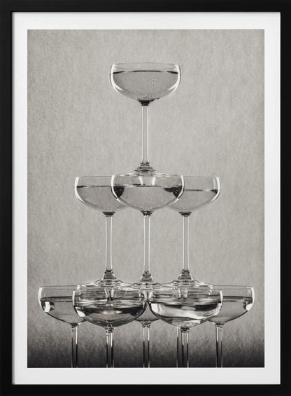 A classic black and white photograph of a champagne tower, with coupe glasses stacked in a pyramid. The glasses are filled with bubbly liquid, and the composition is set against a textured light gray background and enclosed in a black frame. Poster