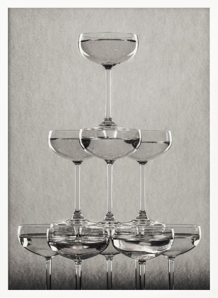 A classic black and white photograph of a champagne tower, with coupe glasses stacked in a pyramid. The glasses are filled with bubbly liquid, and the composition is set against a textured light gray background and enclosed in a black frame. Poster