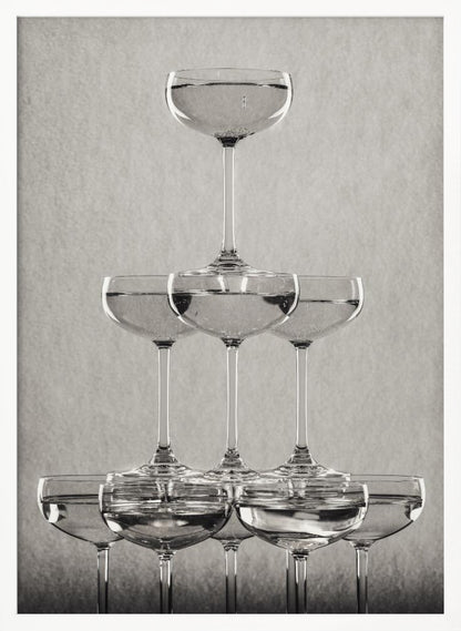 A classic black and white photograph of a champagne tower, with coupe glasses stacked in a pyramid. The glasses are filled with bubbly liquid, and the composition is set against a textured light gray background and enclosed in a black frame. Poster