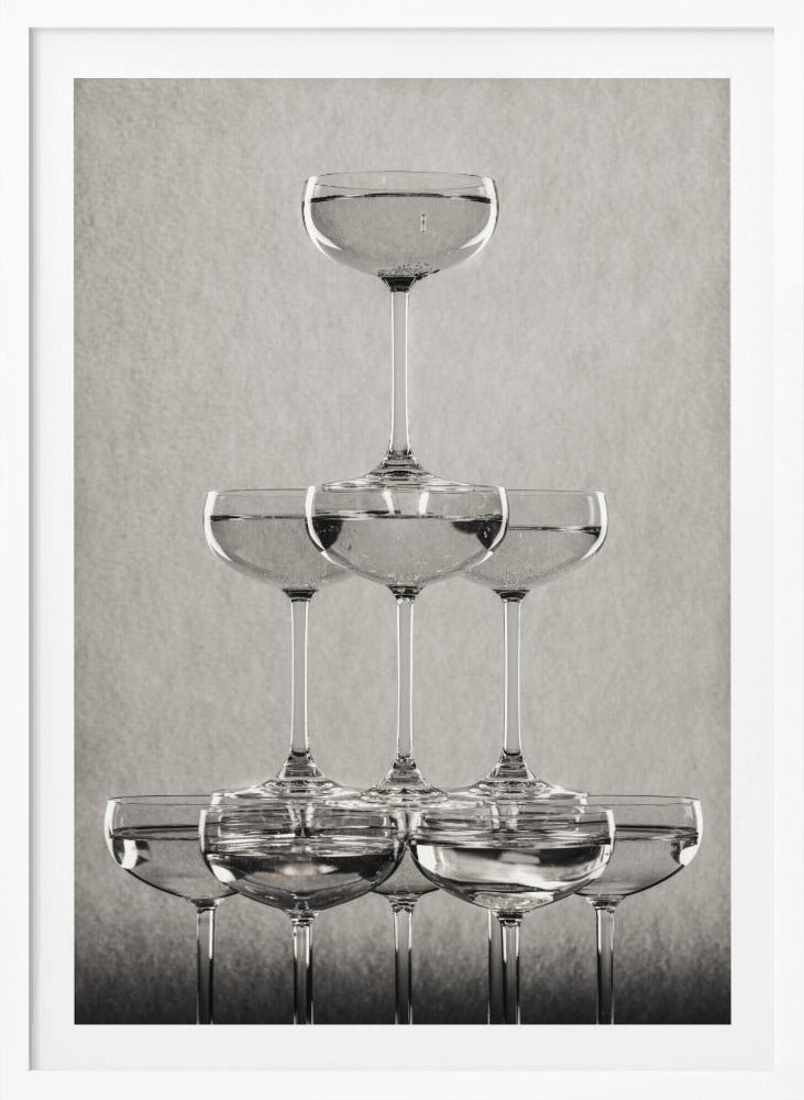 A classic black and white photograph of a champagne tower, with coupe glasses stacked in a pyramid. The glasses are filled with bubbly liquid, and the composition is set against a textured light gray background and enclosed in a black frame. Poster