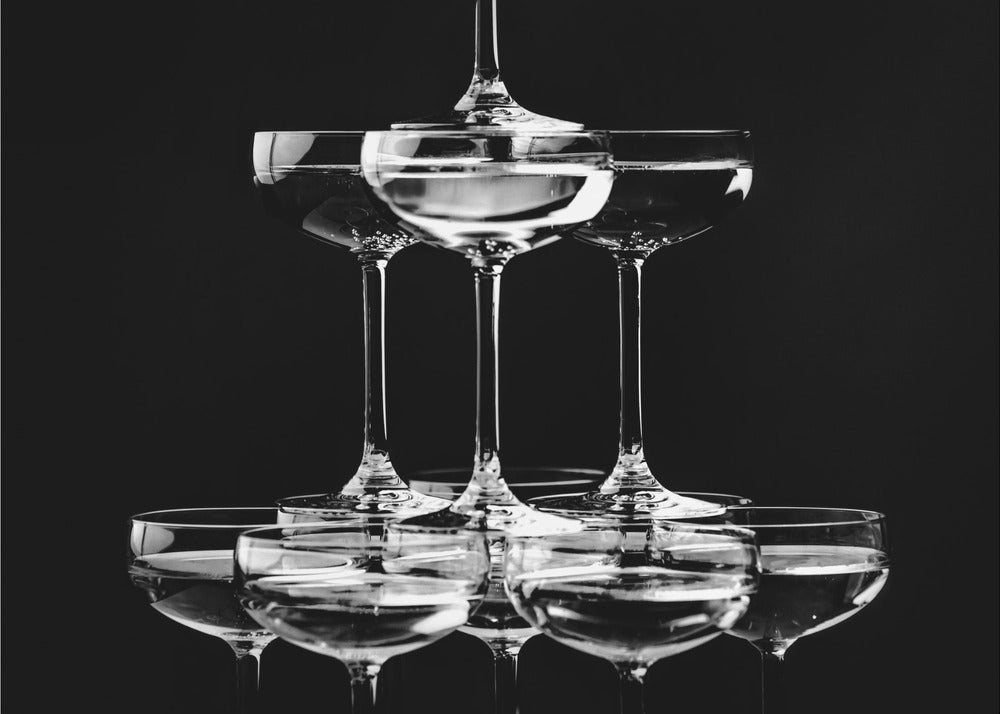 A black and white photograph of a champagne tower made of coupe glasses stacked in a pyramid shape against a solid black background, all within a silver frame. Print