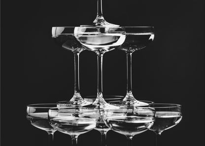 A black and white photograph of a champagne tower made of coupe glasses stacked in a pyramid shape against a solid black background, all within a silver frame. Print