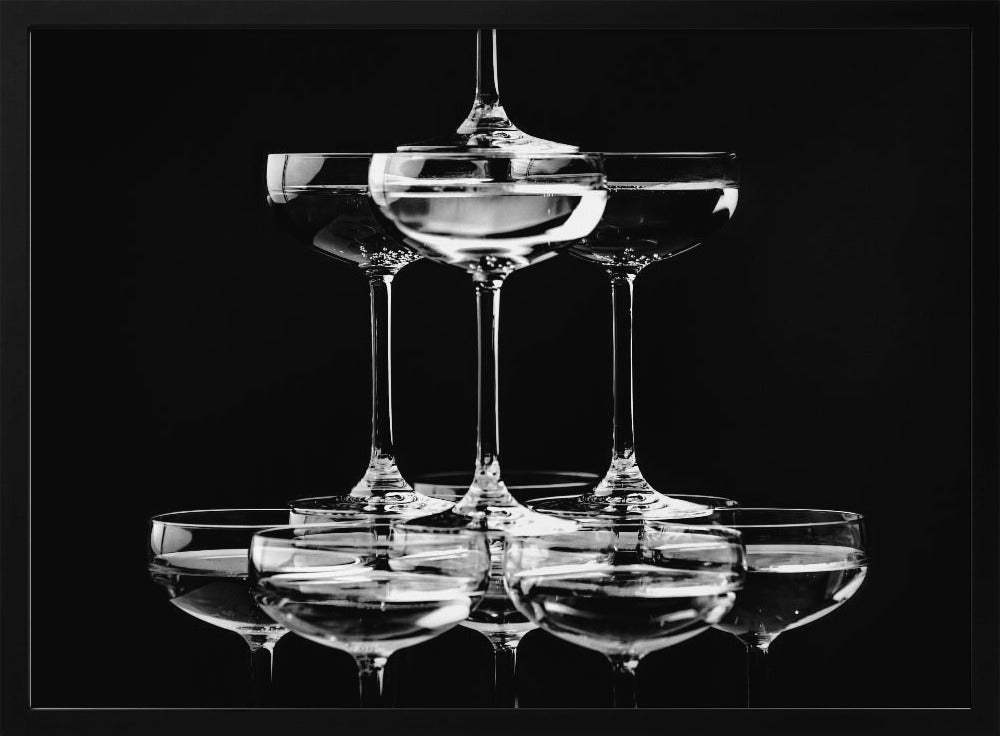 A black and white photograph of a champagne tower made of coupe glasses stacked in a pyramid shape against a solid black background, all within a silver frame. Print