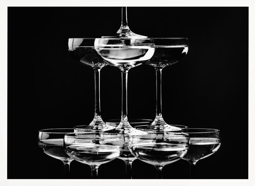 A black and white photograph of a champagne tower made of coupe glasses stacked in a pyramid shape against a solid black background, all within a silver frame. Print