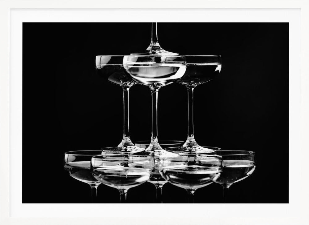A black and white photograph of a champagne tower made of coupe glasses stacked in a pyramid shape against a solid black background, all within a silver frame. Print