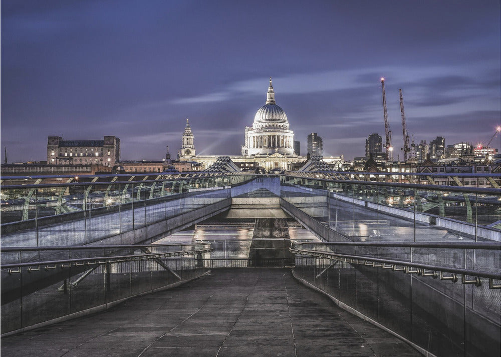 A nighttime photograph from the Millennium Bridge leading towards the brightly illuminated St. Paul's Cathedral against a dark blue twilight sky in London, all enclosed in a silver frame. Poster