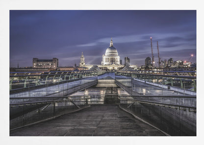 A nighttime photograph from the Millennium Bridge leading towards the brightly illuminated St. Paul's Cathedral against a dark blue twilight sky in London, all enclosed in a silver frame. Poster