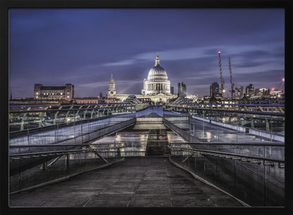 A nighttime photograph from the Millennium Bridge leading towards the brightly illuminated St. Paul's Cathedral against a dark blue twilight sky in London, all enclosed in a silver frame. Poster