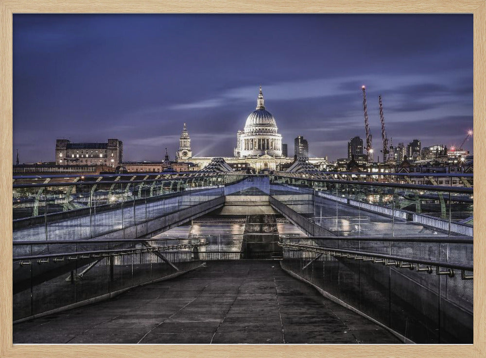 A nighttime photograph from the Millennium Bridge leading towards the brightly illuminated St. Paul's Cathedral against a dark blue twilight sky in London, all enclosed in a silver frame. Poster