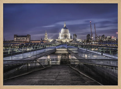 A nighttime photograph from the Millennium Bridge leading towards the brightly illuminated St. Paul's Cathedral against a dark blue twilight sky in London, all enclosed in a silver frame. Poster