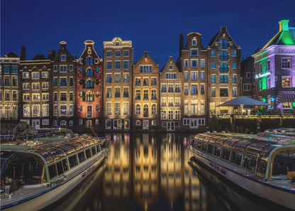 A vibrant night-time photograph of a canal in Amsterdam, flanked by two tour boats in the foreground. Traditional Dutch canal houses are brightly lit, casting warm yellow reflections on the dark water under a deep blue sky. Print