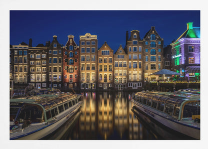 A vibrant night-time photograph of a canal in Amsterdam, flanked by two tour boats in the foreground. Traditional Dutch canal houses are brightly lit, casting warm yellow reflections on the dark water under a deep blue sky. Print