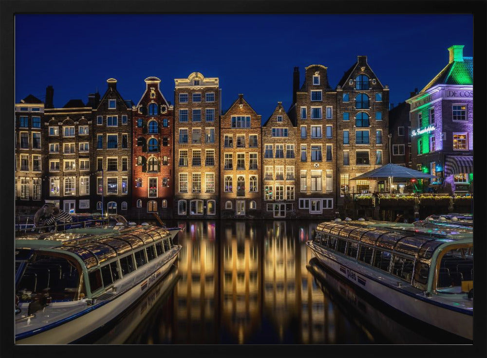 A vibrant night-time photograph of a canal in Amsterdam, flanked by two tour boats in the foreground. Traditional Dutch canal houses are brightly lit, casting warm yellow reflections on the dark water under a deep blue sky. Print