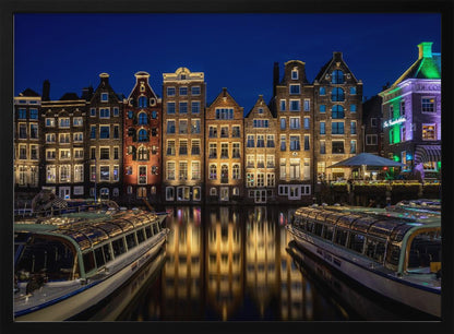A vibrant night-time photograph of a canal in Amsterdam, flanked by two tour boats in the foreground. Traditional Dutch canal houses are brightly lit, casting warm yellow reflections on the dark water under a deep blue sky. Print