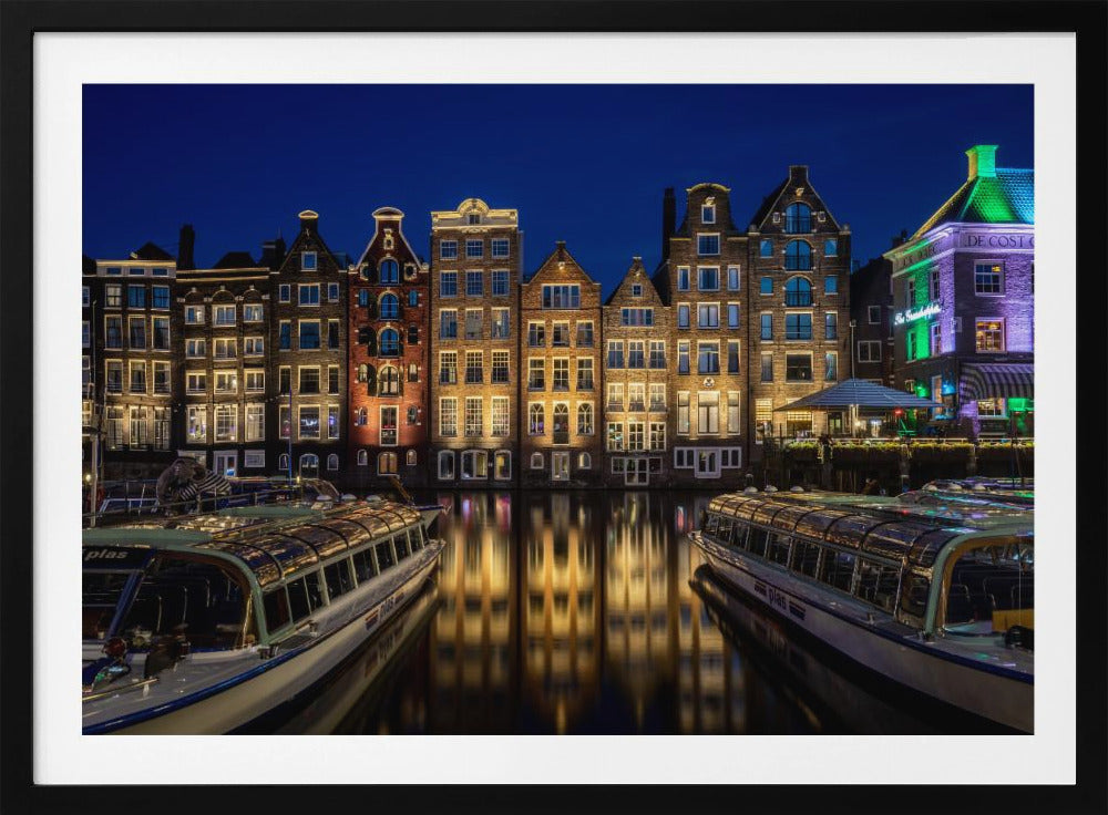 A vibrant night-time photograph of a canal in Amsterdam, flanked by two tour boats in the foreground. Traditional Dutch canal houses are brightly lit, casting warm yellow reflections on the dark water under a deep blue sky. Print