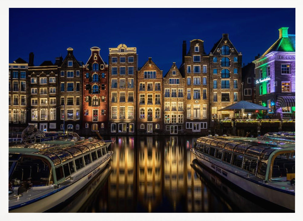 A vibrant night-time photograph of a canal in Amsterdam, flanked by two tour boats in the foreground. Traditional Dutch canal houses are brightly lit, casting warm yellow reflections on the dark water under a deep blue sky. Print