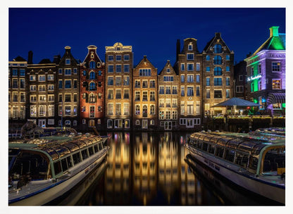 A vibrant night-time photograph of a canal in Amsterdam, flanked by two tour boats in the foreground. Traditional Dutch canal houses are brightly lit, casting warm yellow reflections on the dark water under a deep blue sky. Print