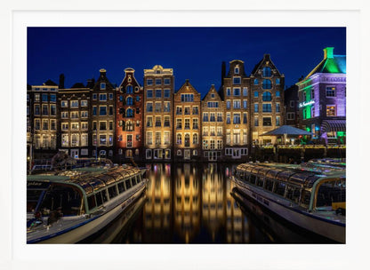 A vibrant night-time photograph of a canal in Amsterdam, flanked by two tour boats in the foreground. Traditional Dutch canal houses are brightly lit, casting warm yellow reflections on the dark water under a deep blue sky. Print