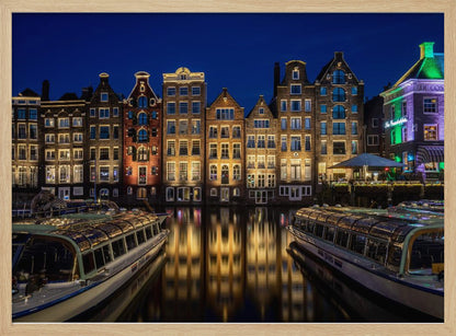 A vibrant night-time photograph of a canal in Amsterdam, flanked by two tour boats in the foreground. Traditional Dutch canal houses are brightly lit, casting warm yellow reflections on the dark water under a deep blue sky. Print
