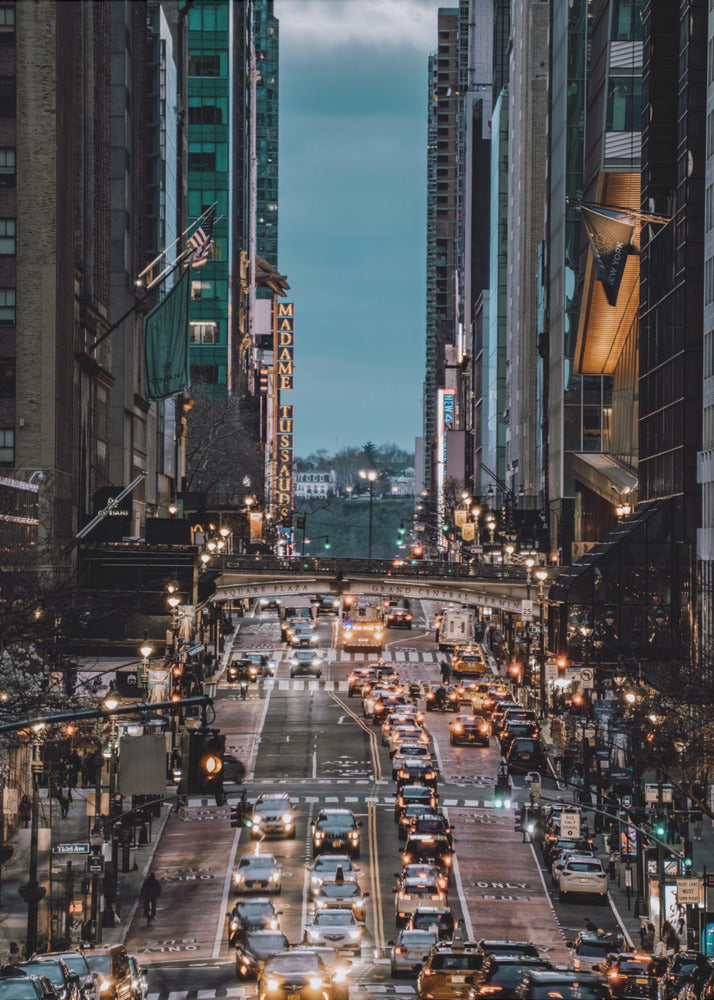 A high-angle, vertical photograph looking down a bustling New York City street at dusk, framed by towering skyscrapers. The street below is packed with cars, their headlights creating a warm golden glow, contrasting with the cool, dark blue tones of the twilight sky and buildings. Poster