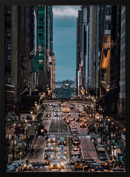 A high-angle, vertical photograph looking down a bustling New York City street at dusk, framed by towering skyscrapers. The street below is packed with cars, their headlights creating a warm golden glow, contrasting with the cool, dark blue tones of the twilight sky and buildings. Poster